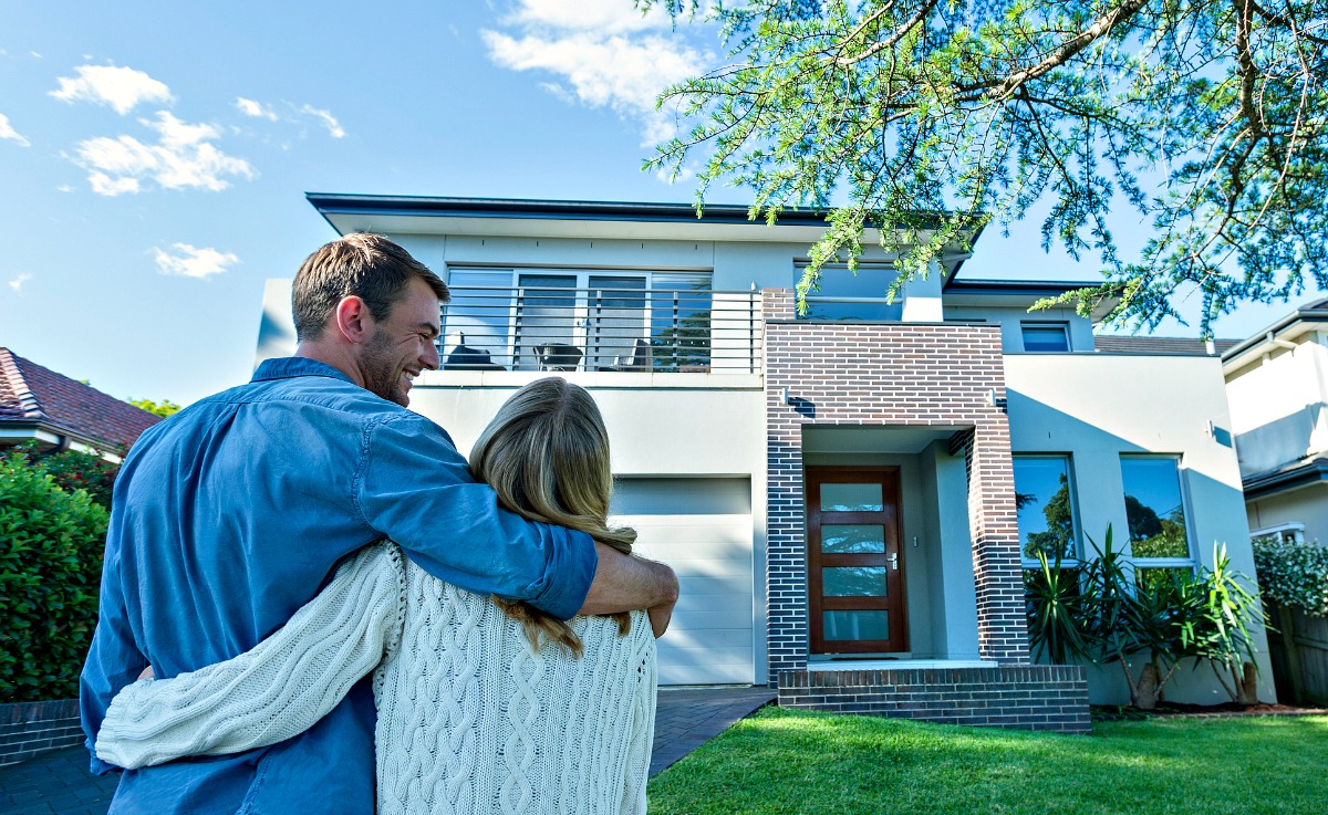 Couple standing in front of their new home. They are both wearing casual clothes and embracing. Rear view from behind them. The house is contemporary with a brick facade, driveway, balcony and a green lawn. The front door is also visible. Copy space (